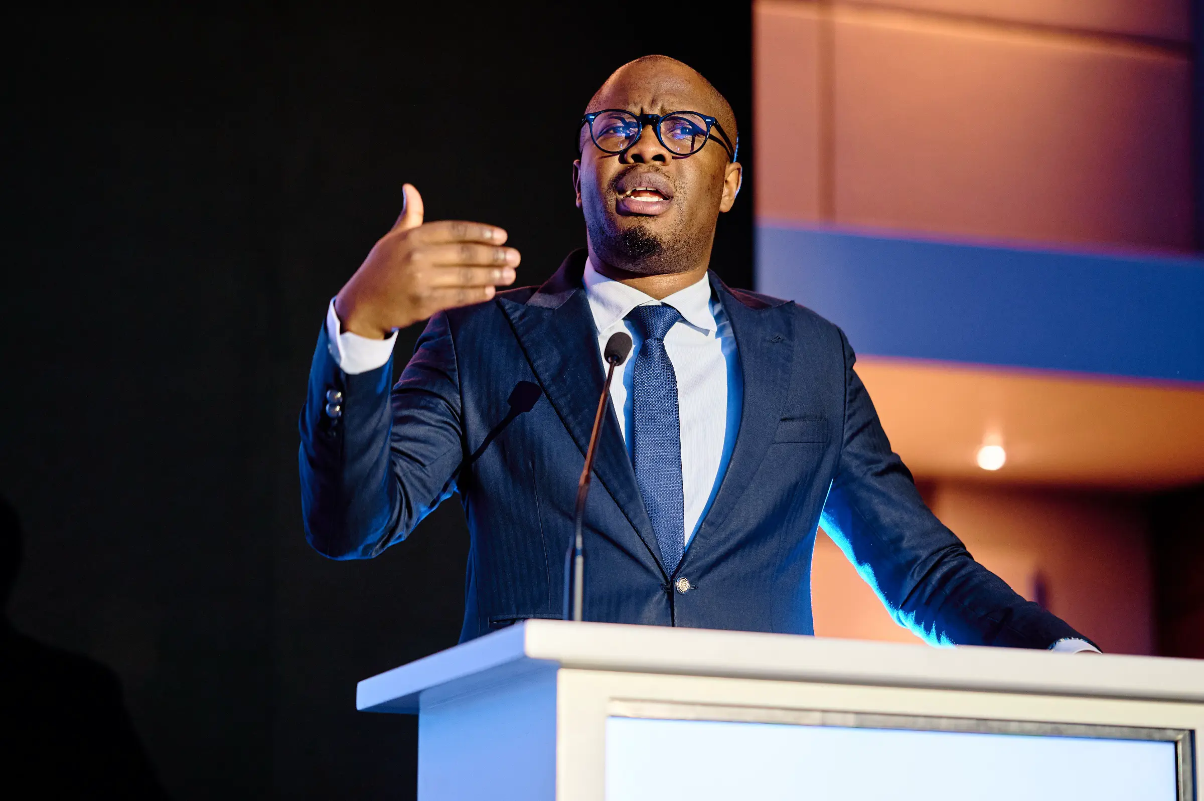 A polished, professional portrait of a conference speaker gesturing during a presentation, capturing dynamic stage lighting.